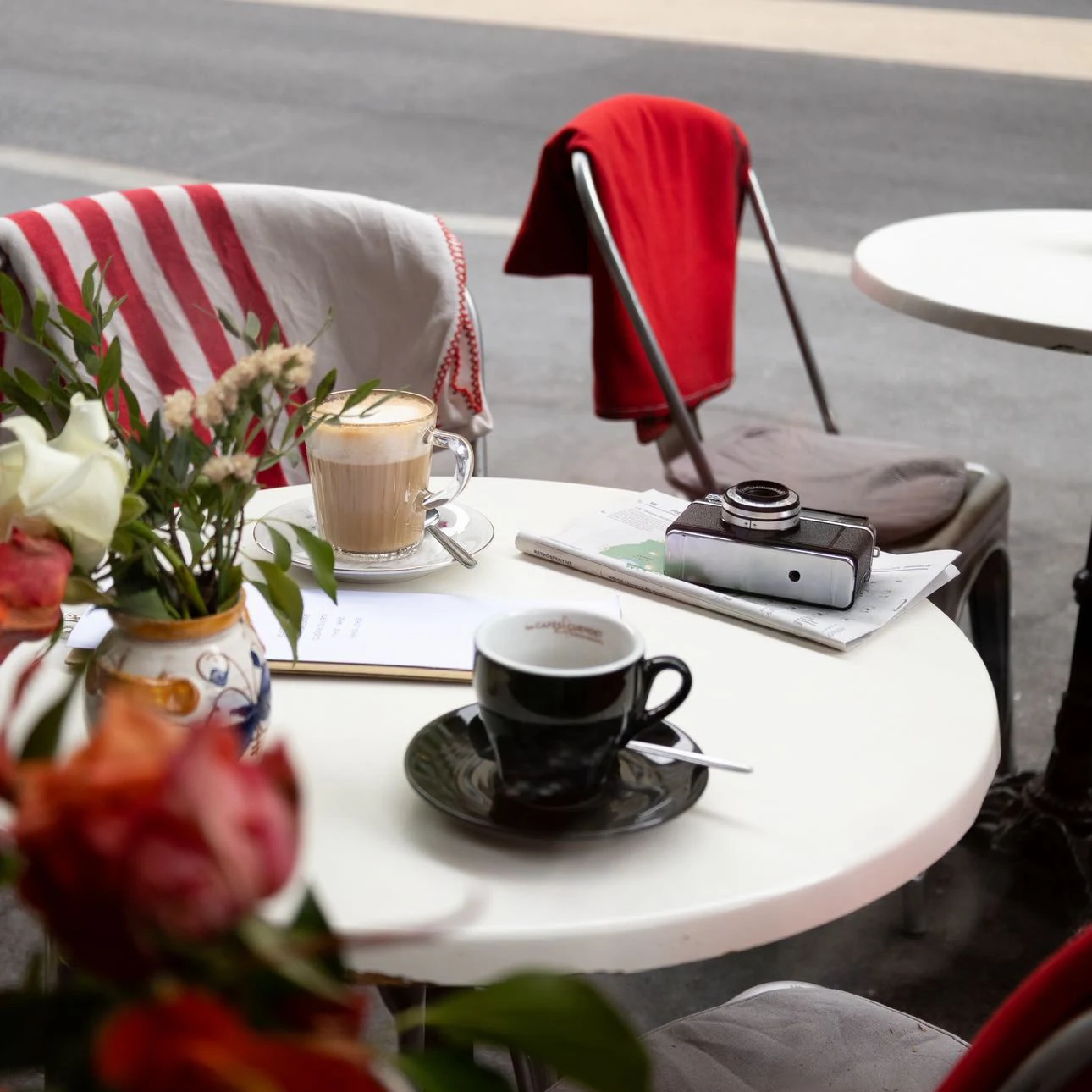 Un latte macchiato et un expresso sur une table ronde en terrasse, à côté d’un appareil photo posé sur un journal et d’un bouquet dans un pot en faïence portugaise.