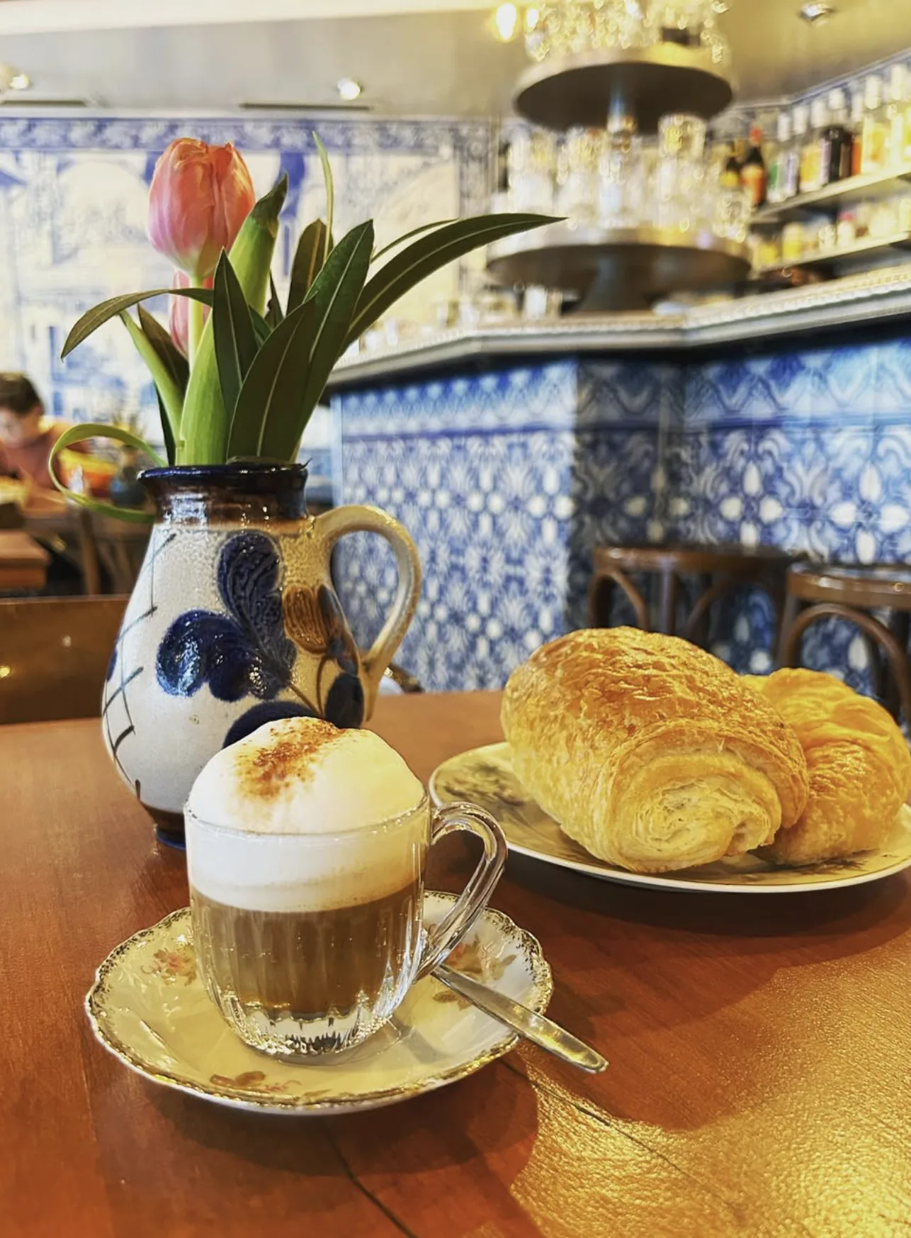 Un cappuccino et deux croissants sur une table du café ; derrière, le comptoir et les grandes fresques d’azulejos bleu et blanc.