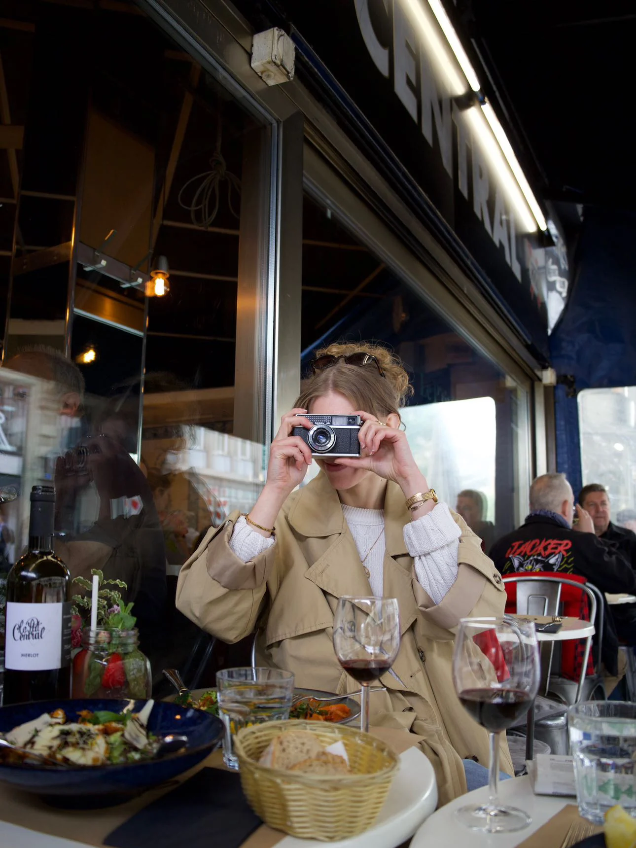 Une cliente, appareil photo à la main, à une table en terrasse sous l’enseigne CENTRAL ; sur la table, une bouteille de merlot de la maison, une assiette et une corbeille de pain.