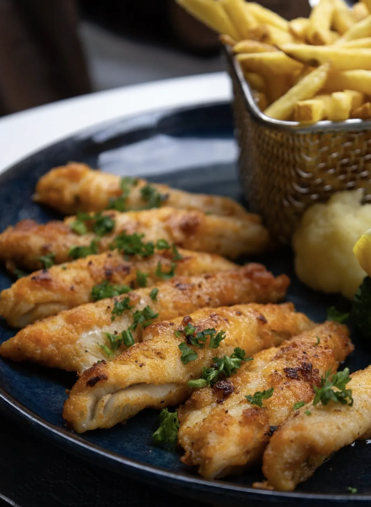 Filets de perche dorés au beurre et au persil sur une assiette bleue, avec un panier de frites et des légumes.
