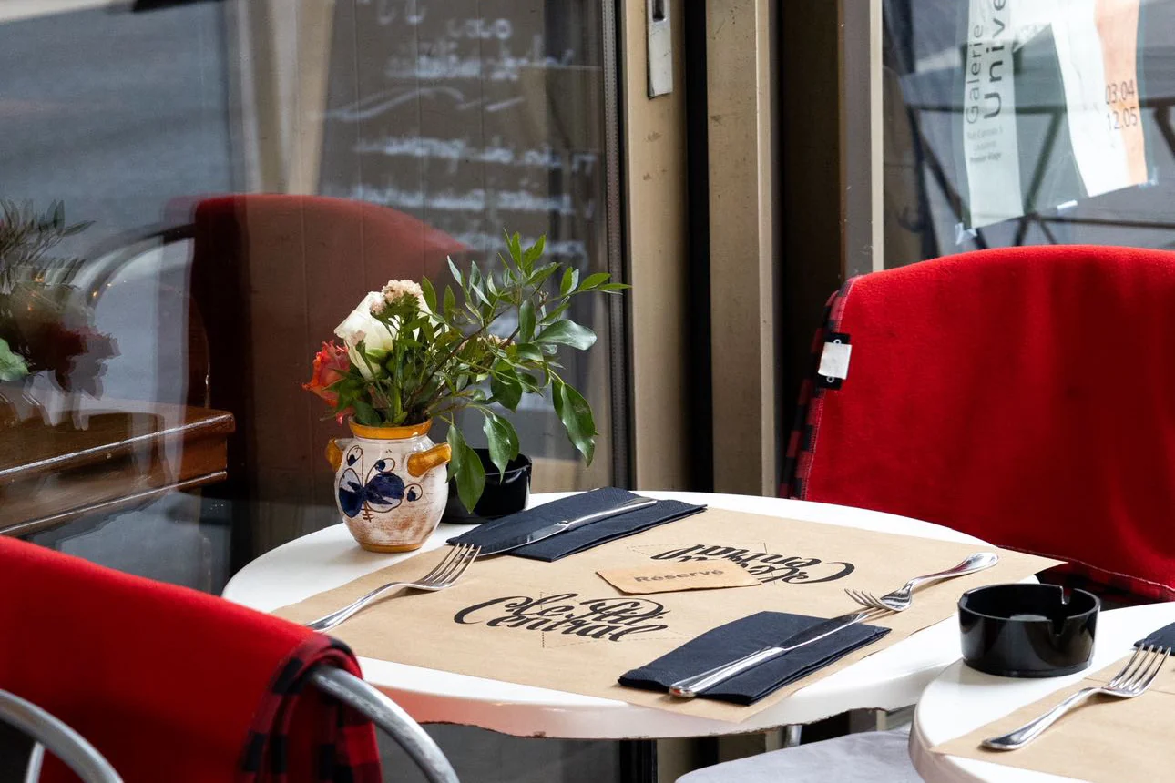 Une table ronde dressée sur un set Le P’tit Central : couverts, serviettes bleues, carton de réservation et un bouquet dans un pot en faïence portugaise, entre deux chaises de velours rouge.