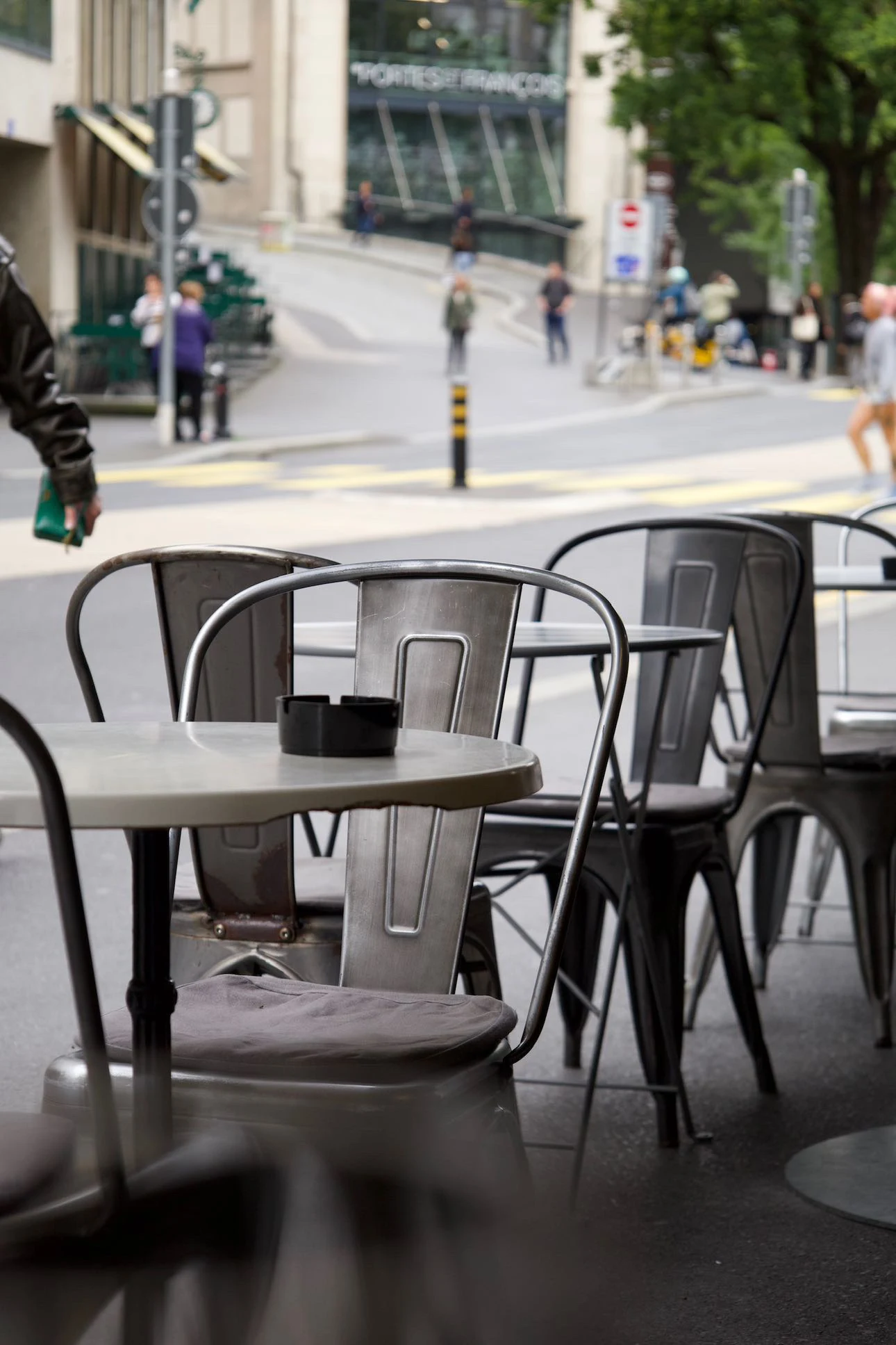Les tables et les chaises en métal de la terrasse, vides avant le service ; derrière, la rue Centrale et les passants.