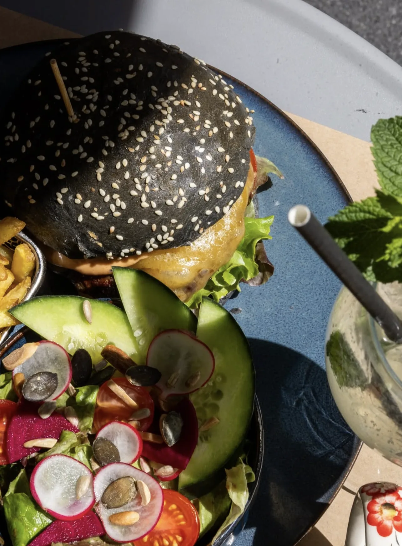 Un burger au pain noir, une salade de saison et un mojito sur une table en terrasse, en plein soleil.
