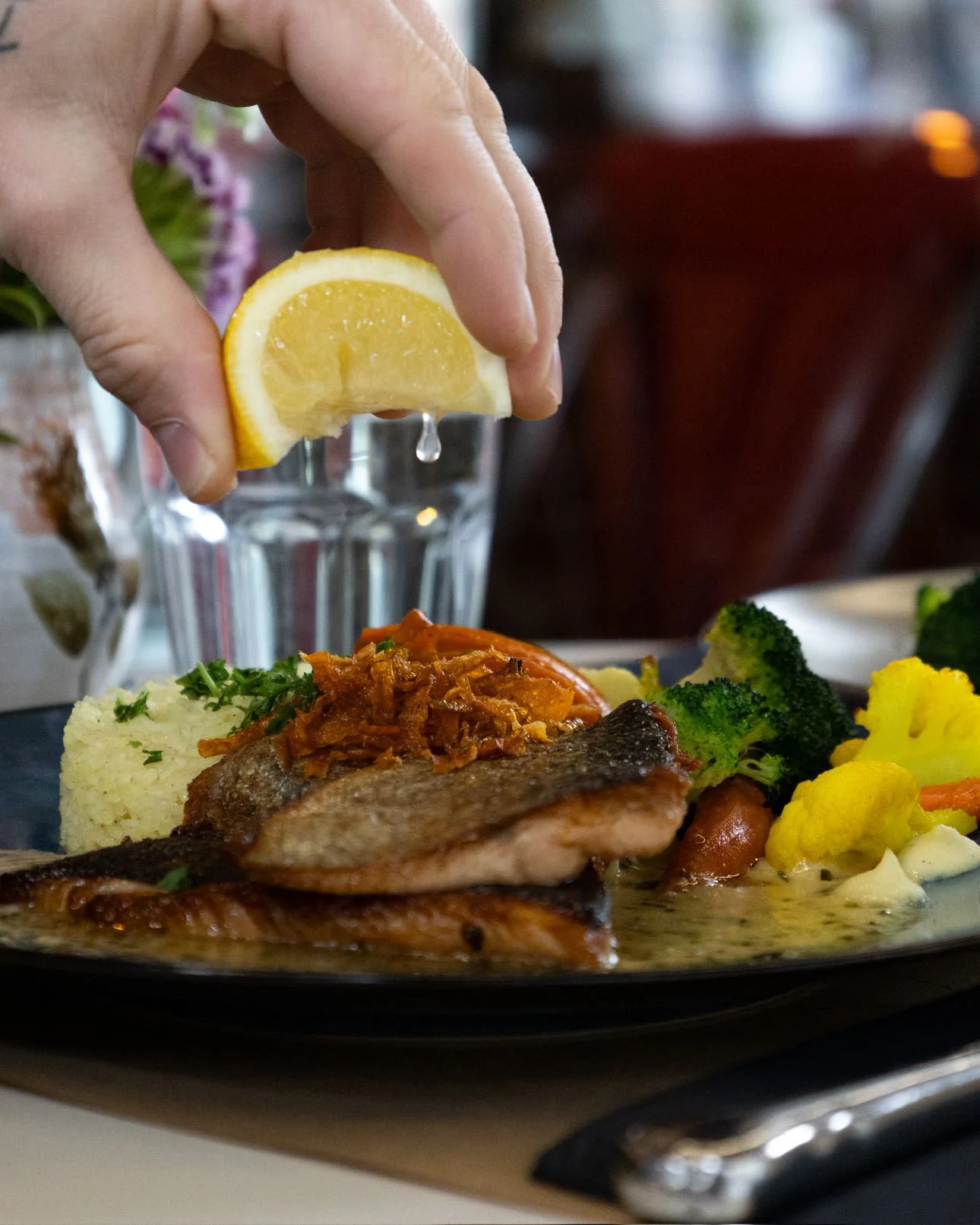 Une main presse un quartier de citron au-dessus d’un filet de truite saumonée snacké, servi avec semoule, oignons frits, brocolis et légumes d’été.