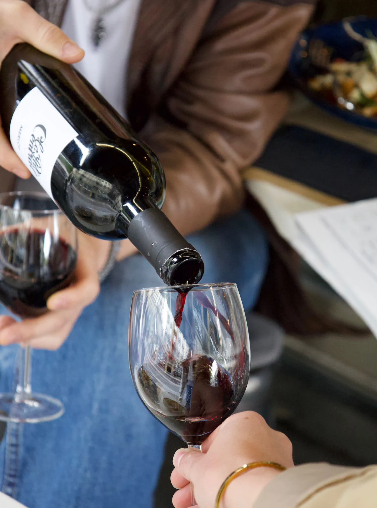 Une bouteille du merlot de la maison versée dans un verre de vin rouge tenu à table ; un second verre déjà servi attend à côté.